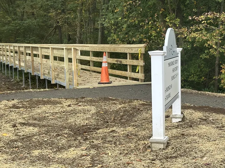 Wooden bridge under construction with a traffic cone. Nearby, a sign stands on a grassy area. Trees are in the background.