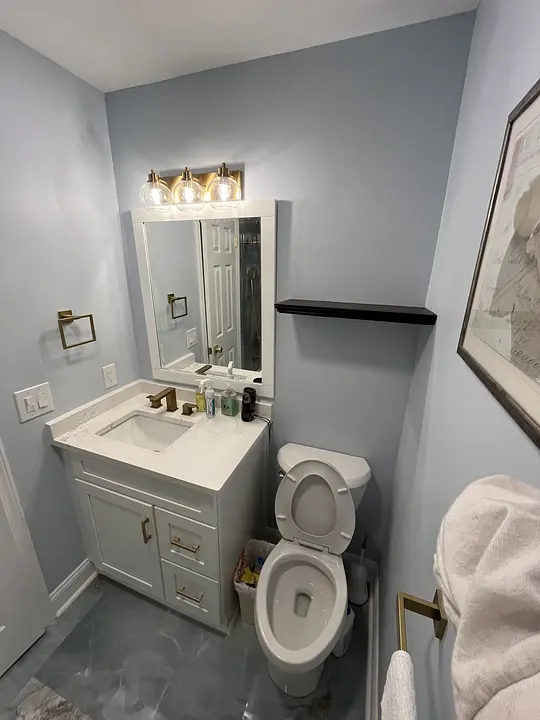 Modern bathroom with white vanity, sink, toilet, and mirror. Three lights above. Blue-gray walls and gold fixtures.