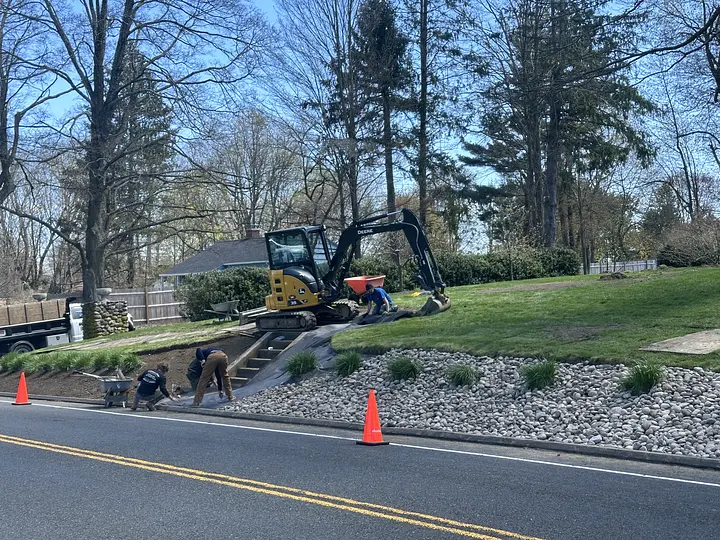 Workers using excavator and wheelbarrow for landscaping beside a road, with trees and a house in the background.