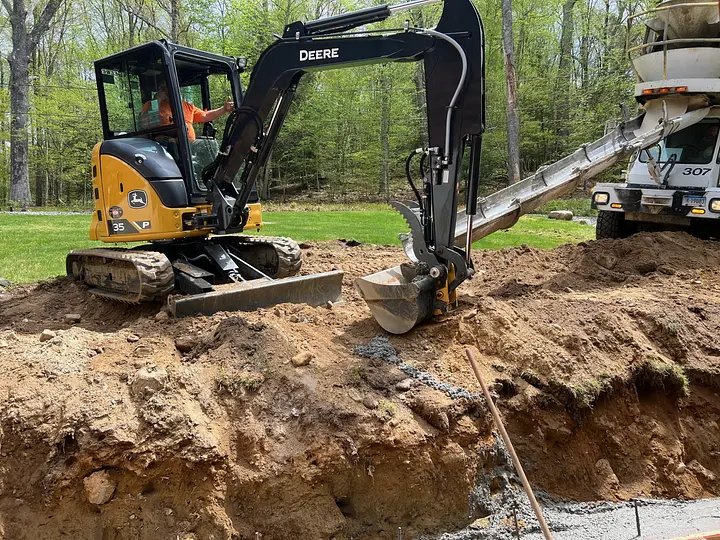 Excavator operator digs soil as concrete flows from a mixer truck chute into a foundation, surrounded by trees.