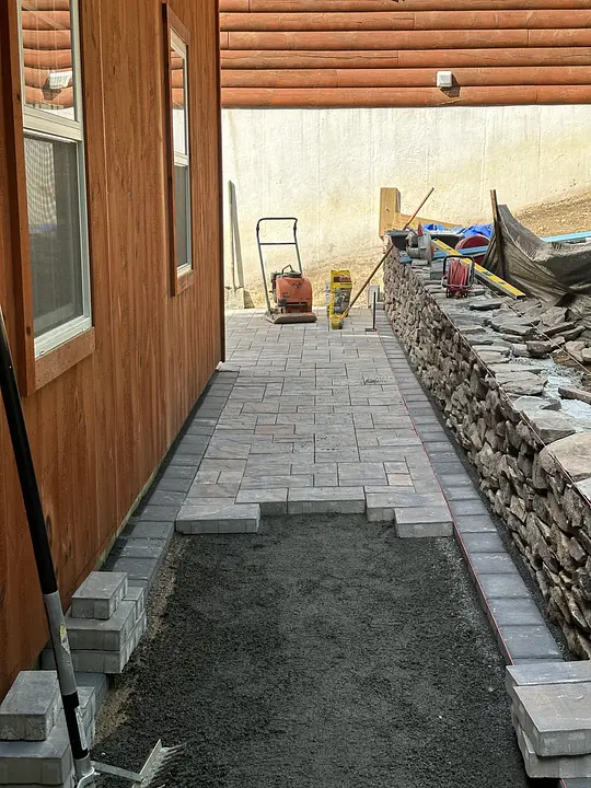 Brick patio under construction beside a wooden building and stone wall, with tools and equipment visible.